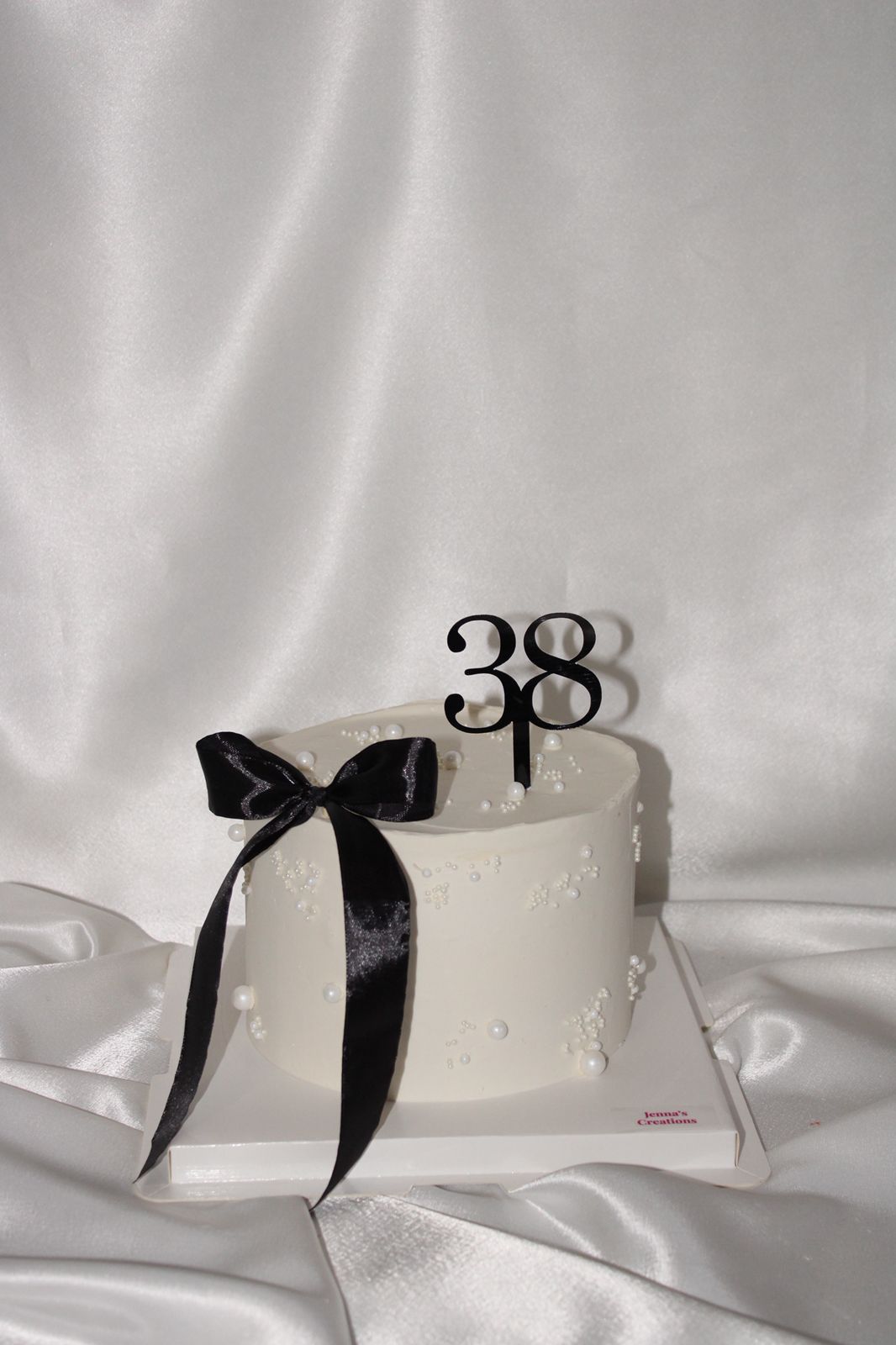 A white frosted birthday cake with a black satin bow and black '38' number topper, displayed on a white board against a draped white satin fabric background.