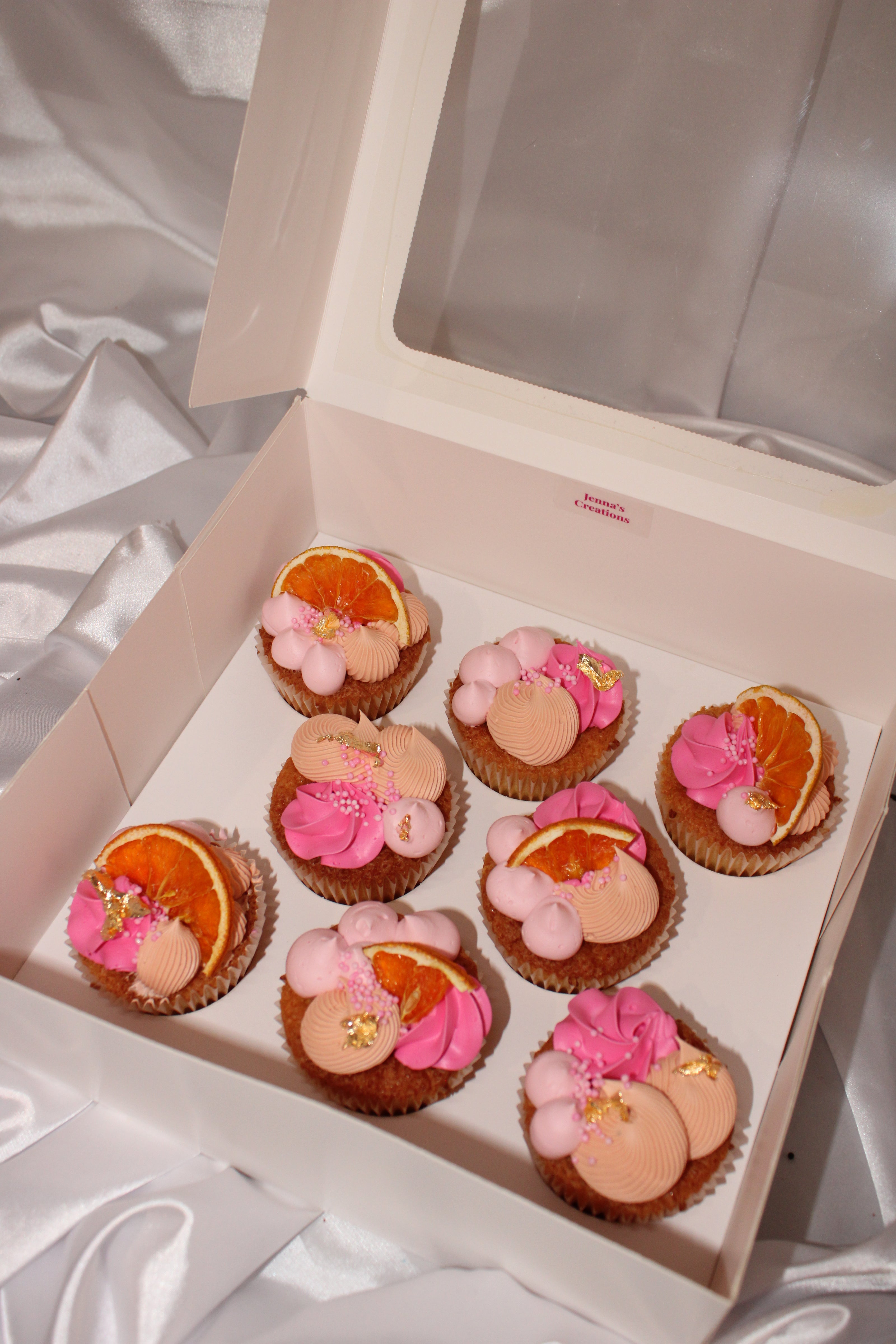 A white bakery box with a clear lid containing nine decorated cupcakes arranged in two rows, set against a draped white satin fabric background.