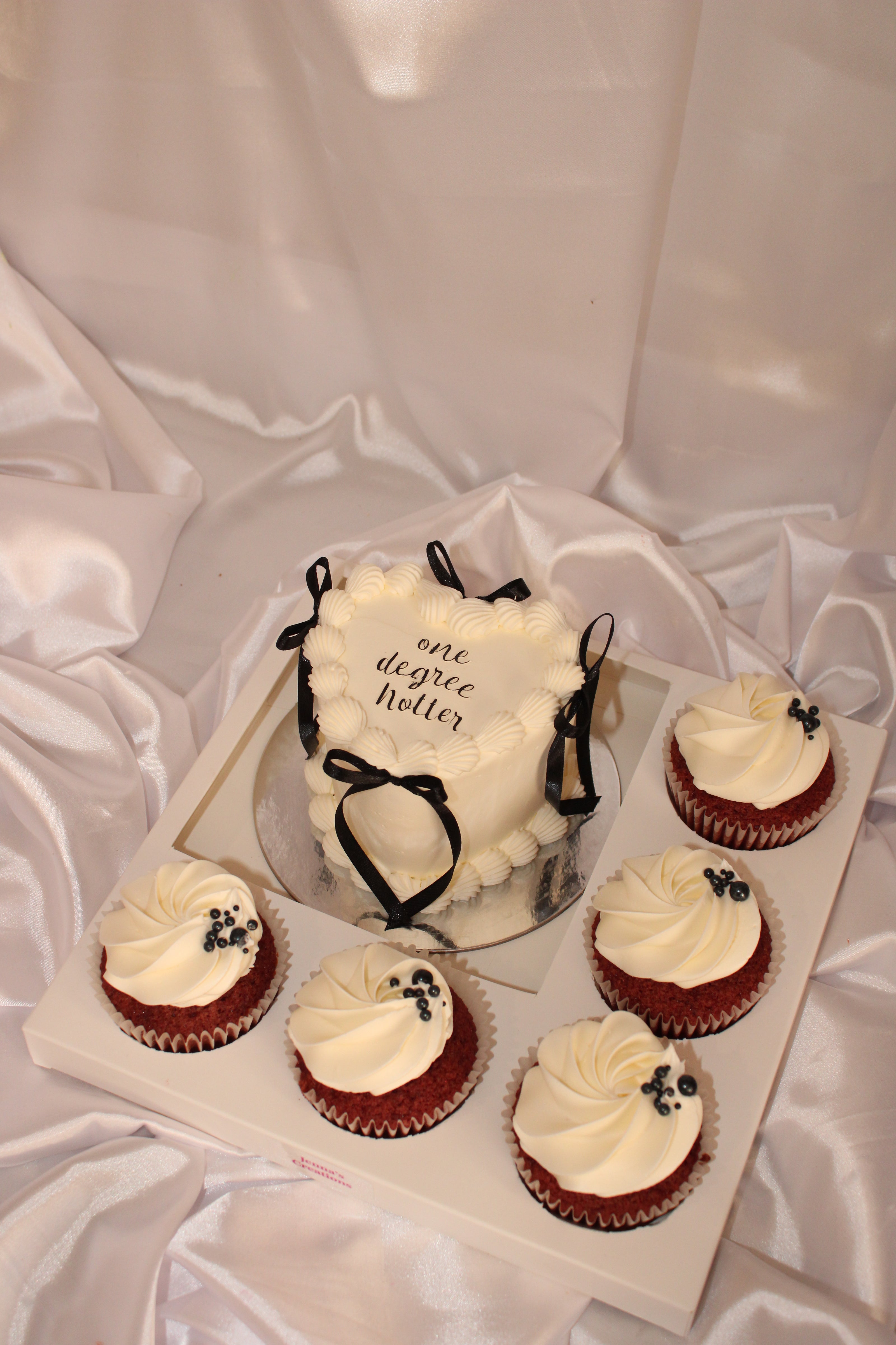 A white heart-shaped frosted cake with black ribbon bows and the words 'one degree hotter' on top, displayed with six matching cupcakes on a white tray against a draped white satin fabric background.