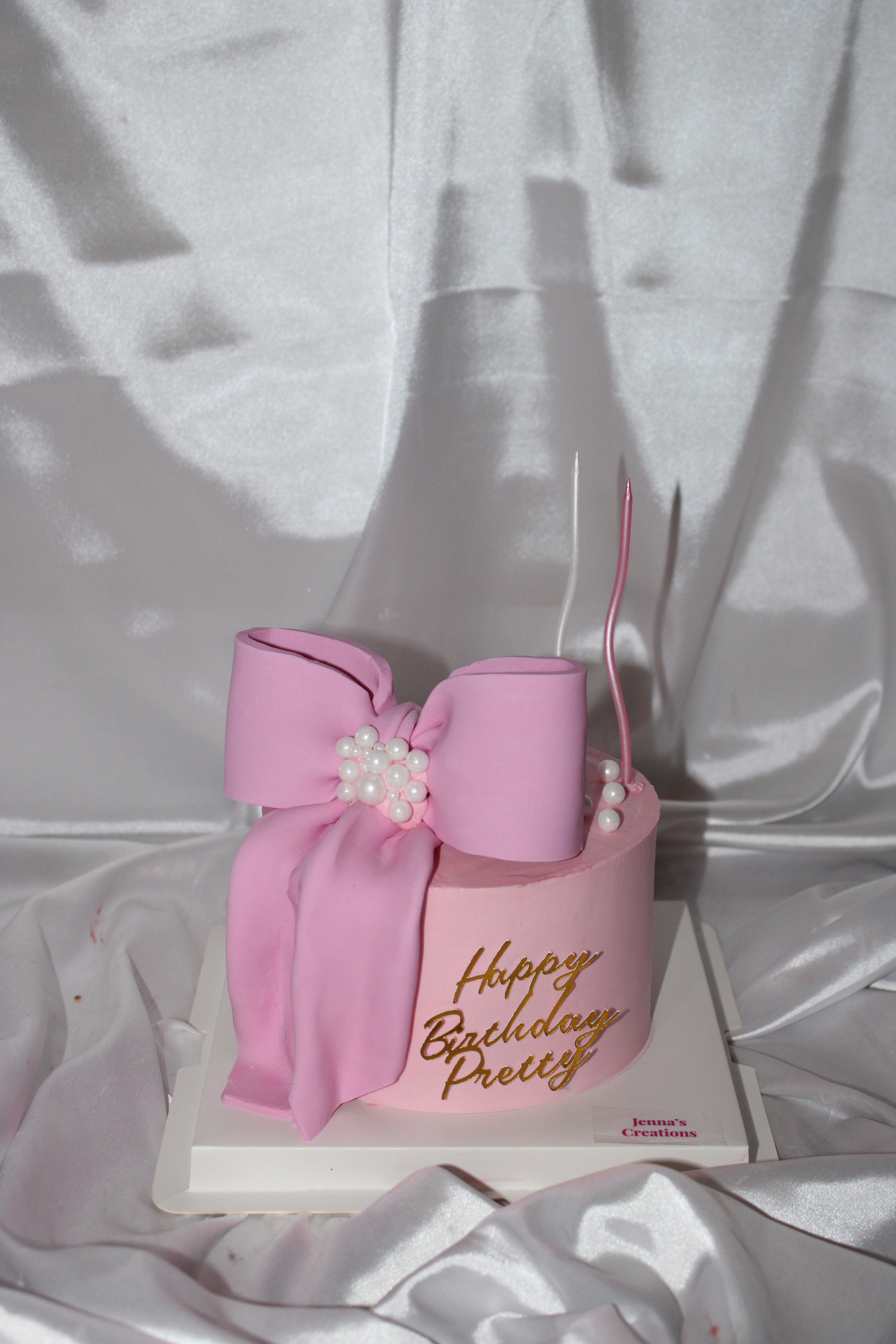 Pink birthday cake with a bow and 'Happy Birthday Pretty' text on a white fabric background