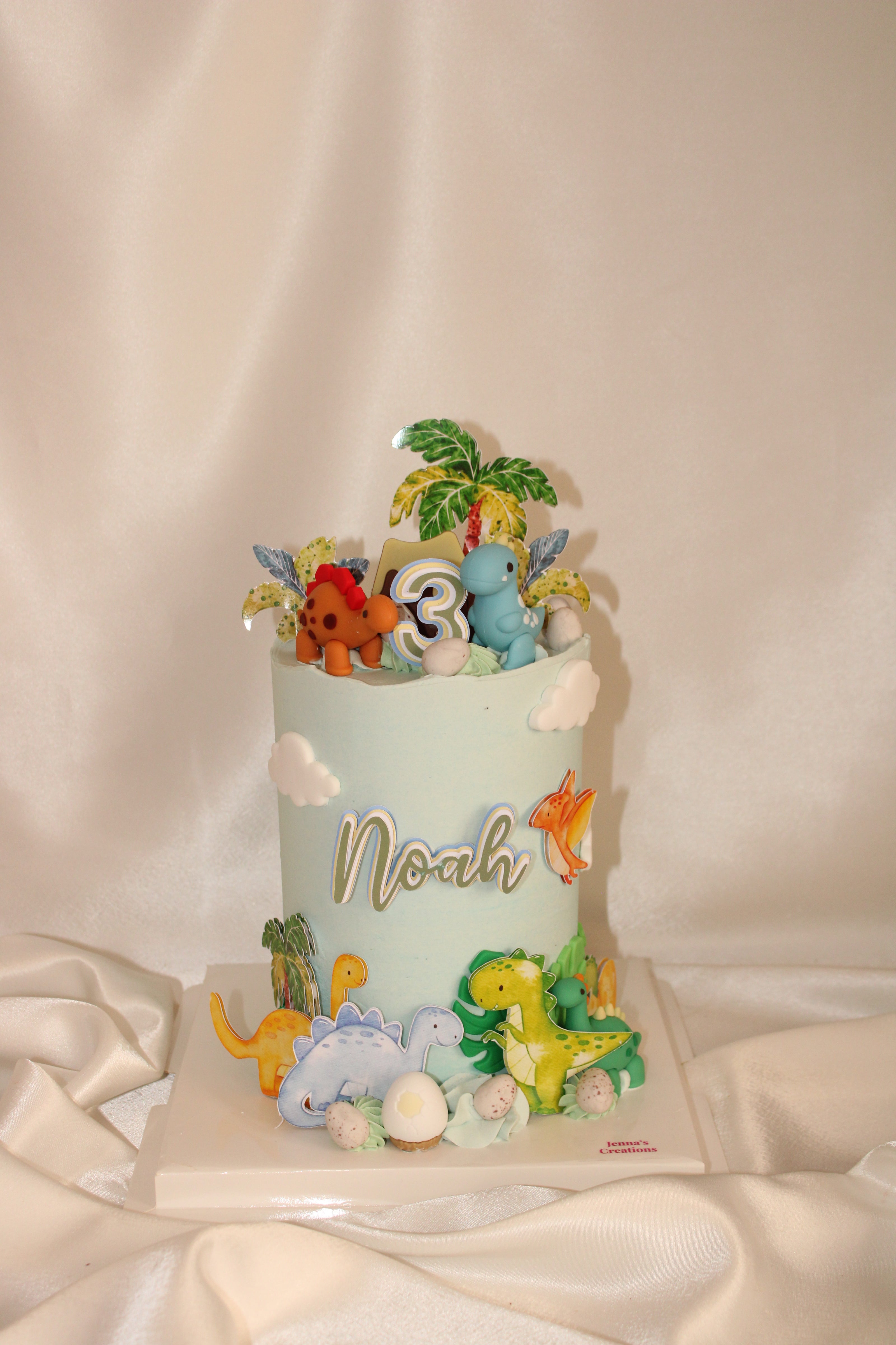 A custom dinosaur-themed birthday cake with a pale blue body, decorated with various colorful fondant dinosaurs, a small palm tree, and the name 'Noah' in gold script on the front, set against a draped cream fabric background.