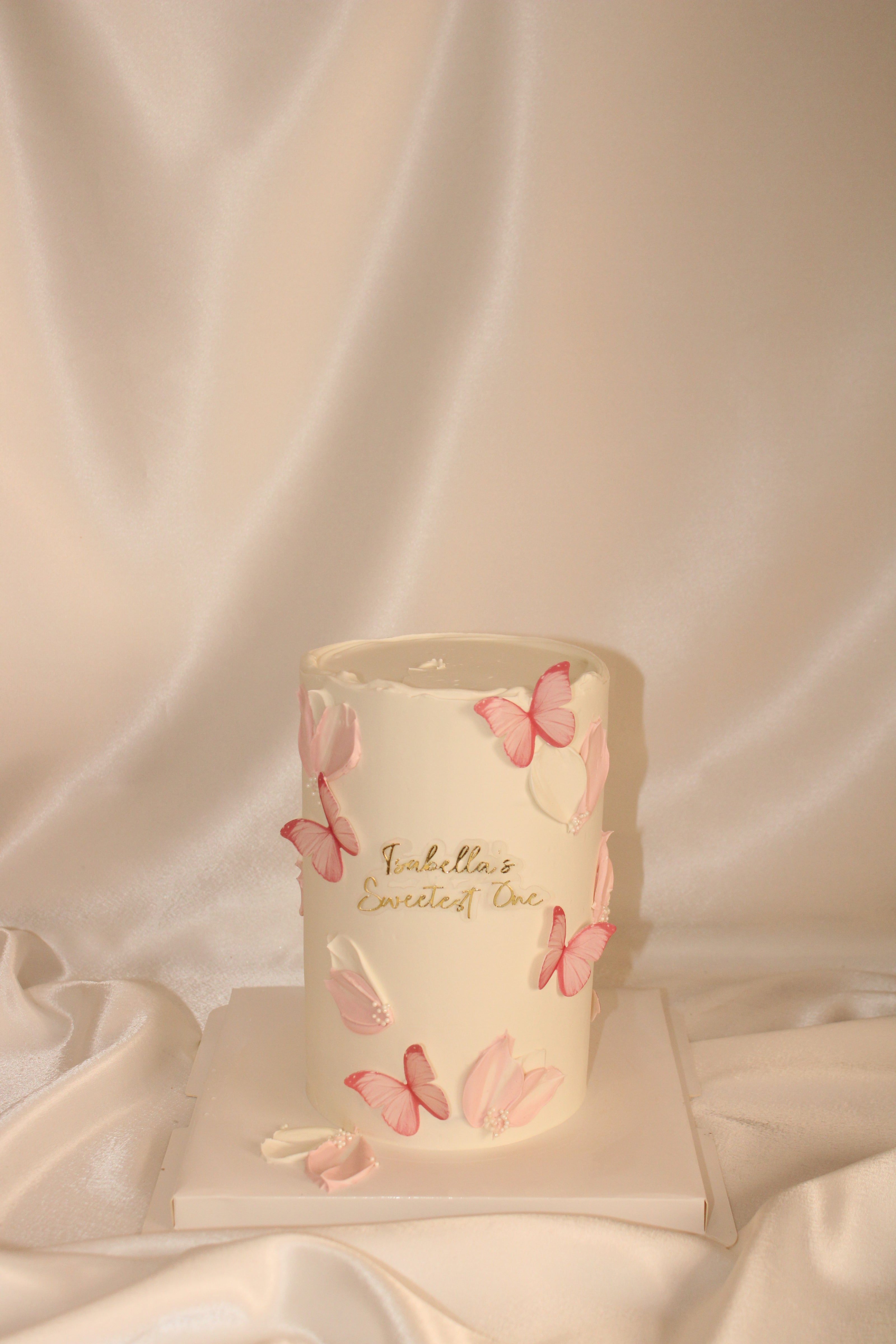 A white cylindrical candle decorated with pink and white butterfly motifs and gold script reading 'Tibetan's Everyday Love', displayed on a white pedestal against a draped satin fabric background.