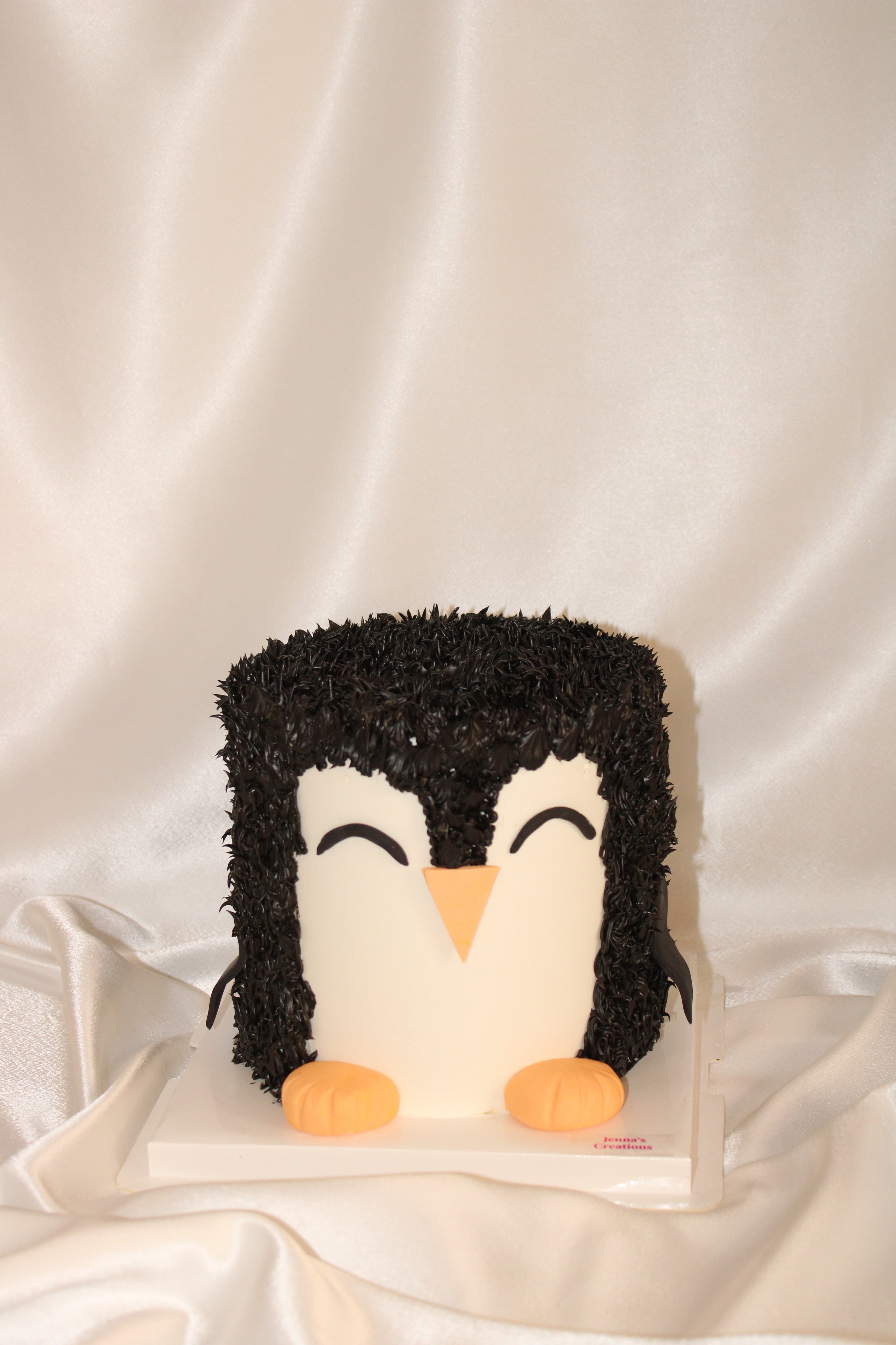 A penguin-shaped cake decorated with black and white frosting, featuring orange feet and beak, displayed on a white base against a draped white satin fabric background.