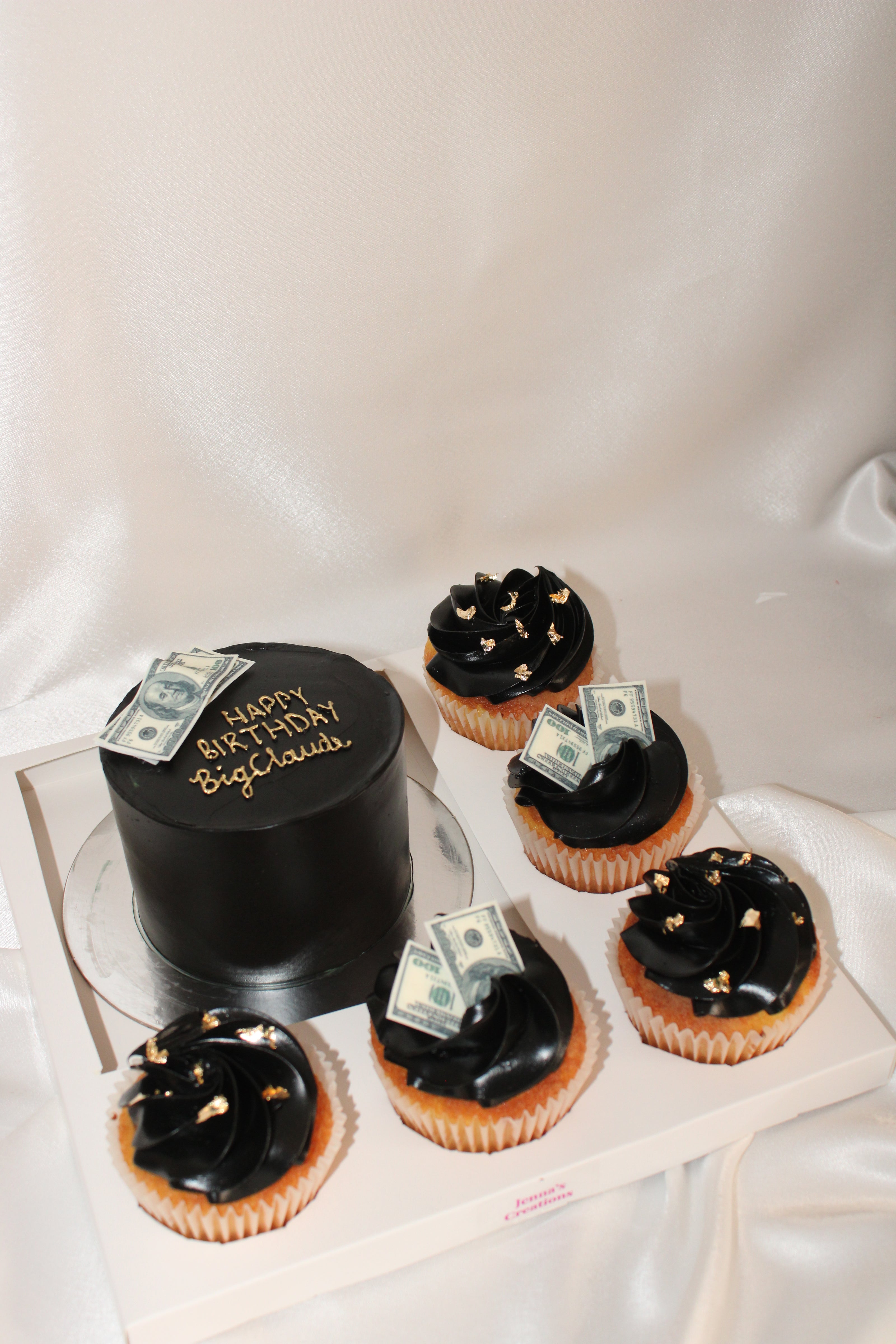 A black frosted birthday cake and five matching black-frosted cupcakes arranged on a white tray against a draped satin fabric background. The cake has gold lettering reading 'HAPPY BIRTHDAY Bigbucks' and small dollar bill decorations on top.