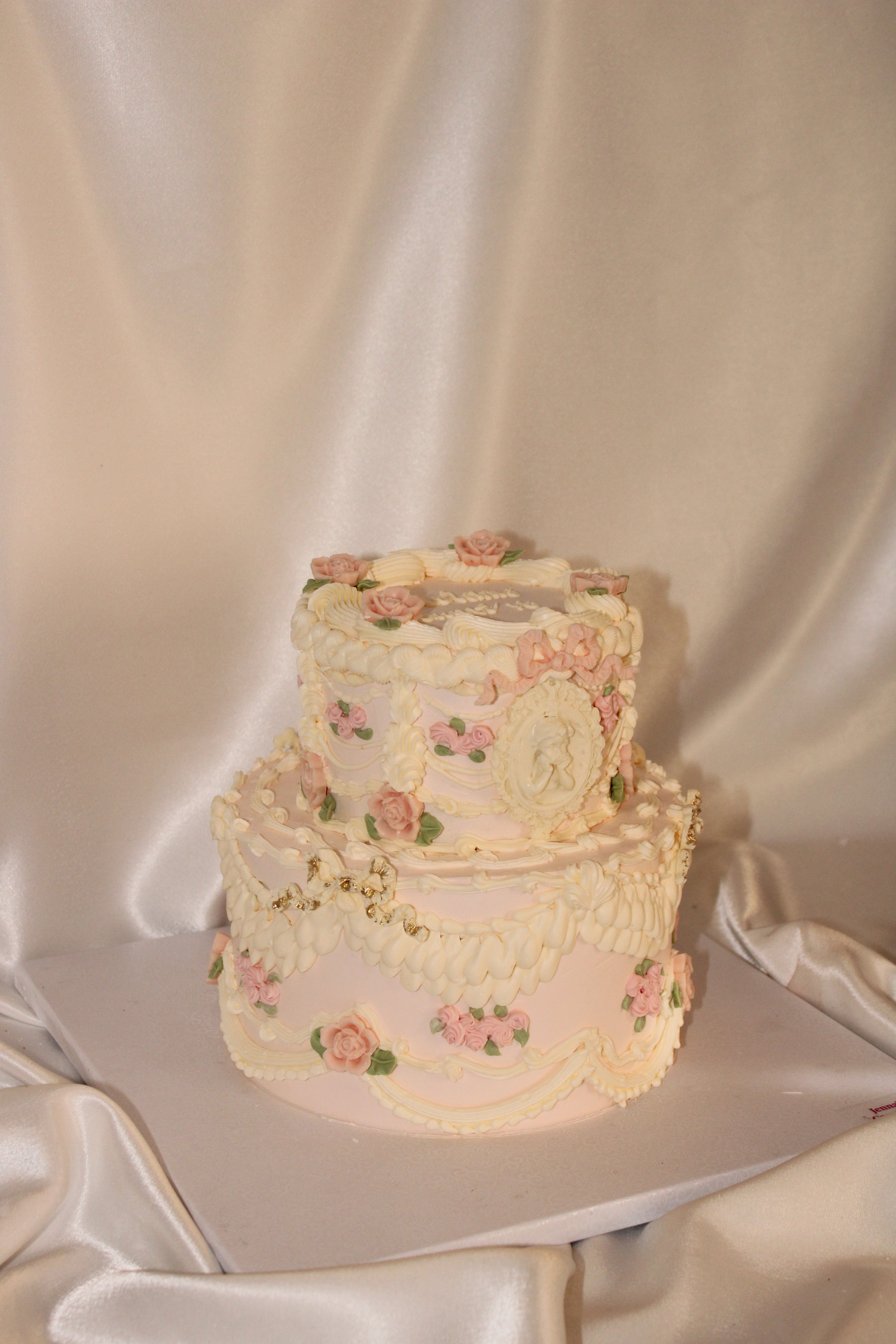 A two-tiered decorated cake with pale pink and white frosting, featuring delicate floral details, gold accents, and a central oval plaque, set against a draped satin fabric backdrop.