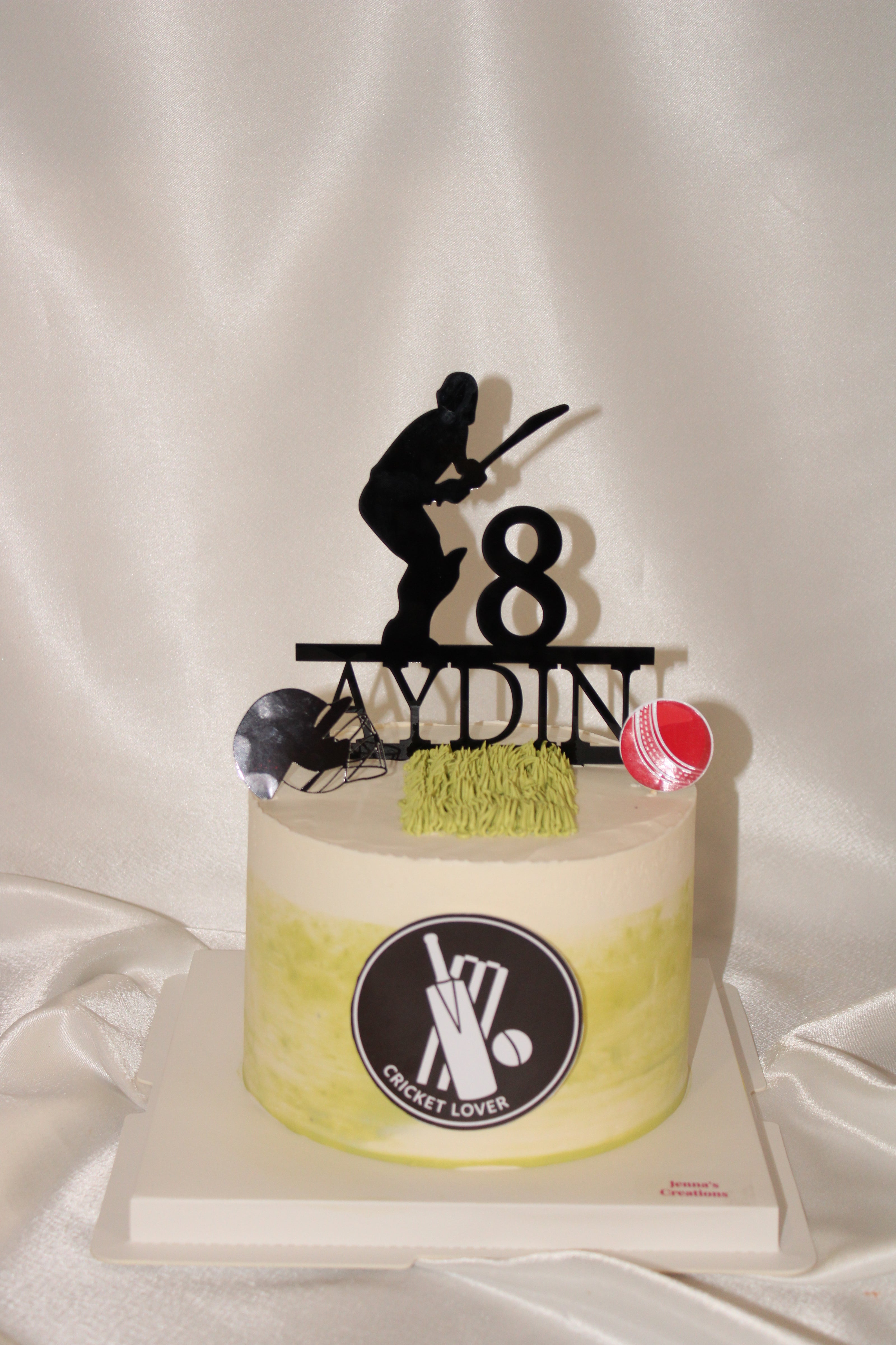 A custom round cake decorated with a yellow-green gradient, featuring a black circular 'Cricket Lover' logo on the front. The cake is topped with a black silhouette of a cricket player, the number 8, the name 'AYDIN', a small green grass-like decoration, and two small cricket-themed decorations.