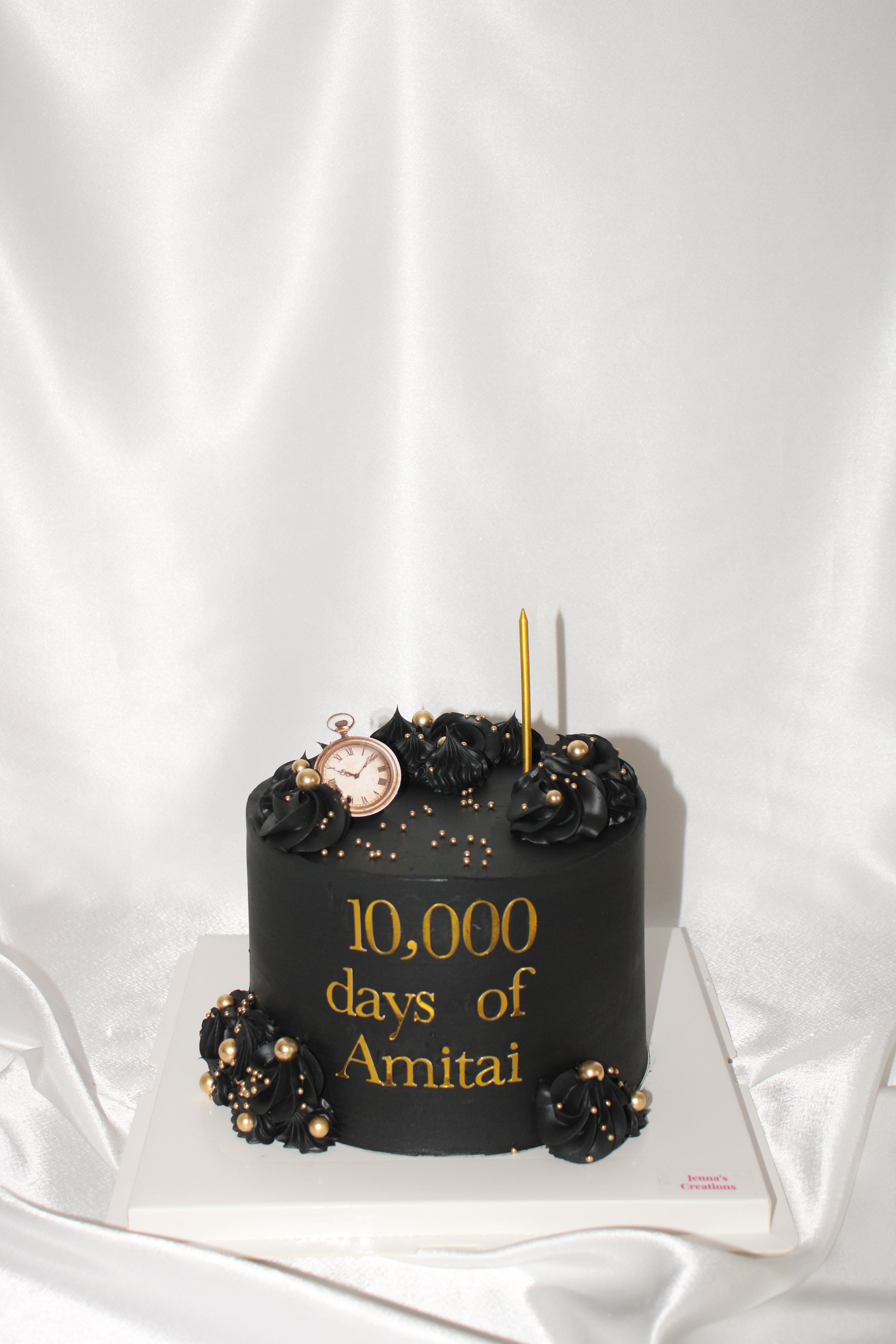 A black cylindrical cake decorated with black roses, gold confetti, a small gold pocket watch, and gold text reading '10,000 days of Amitai' on a white satin backdrop.