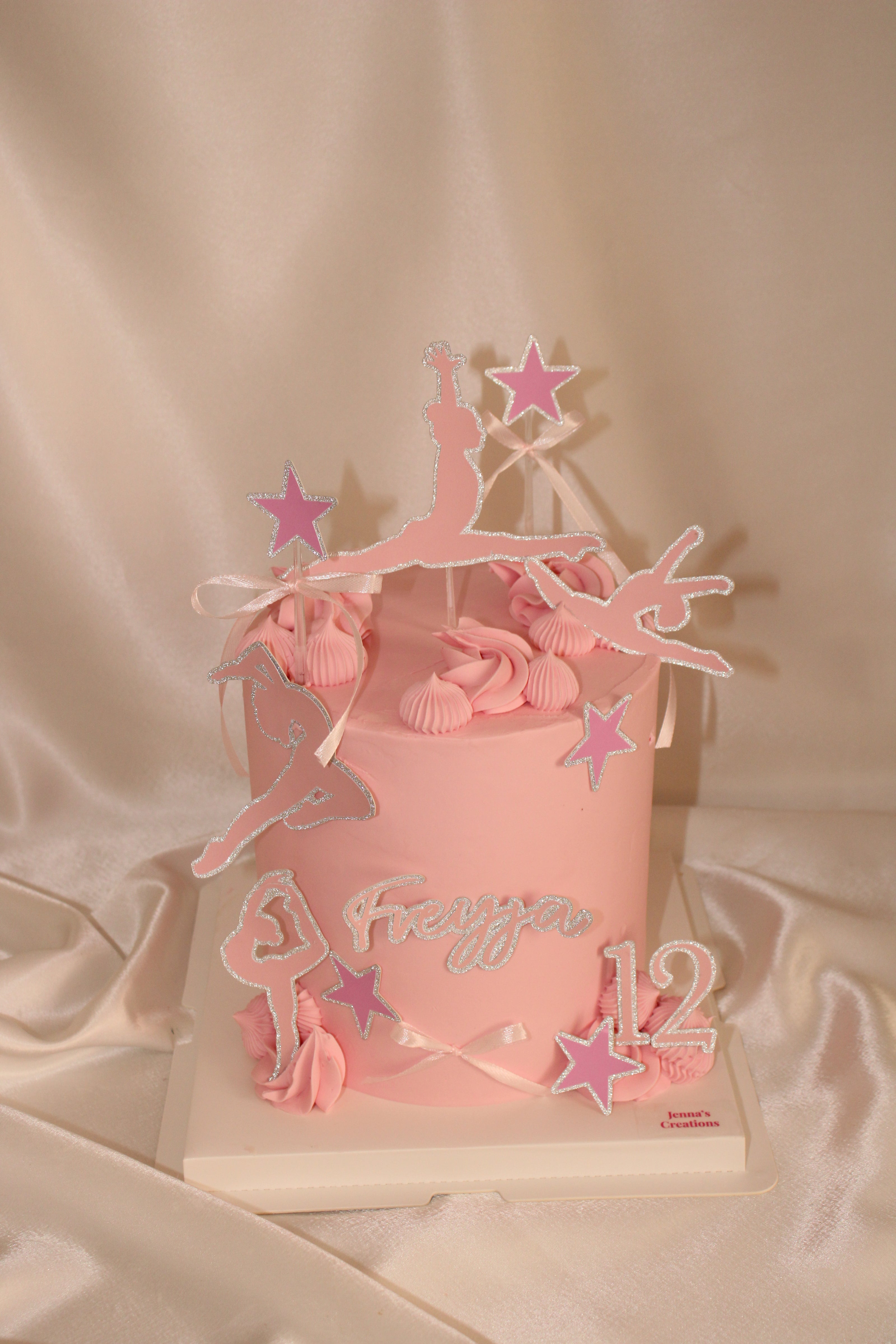 A pink birthday cake decorated with ballerina silhouettes, pink stars, and the name 'Freya' and number '12' in white icing, set on a satin fabric backdrop.