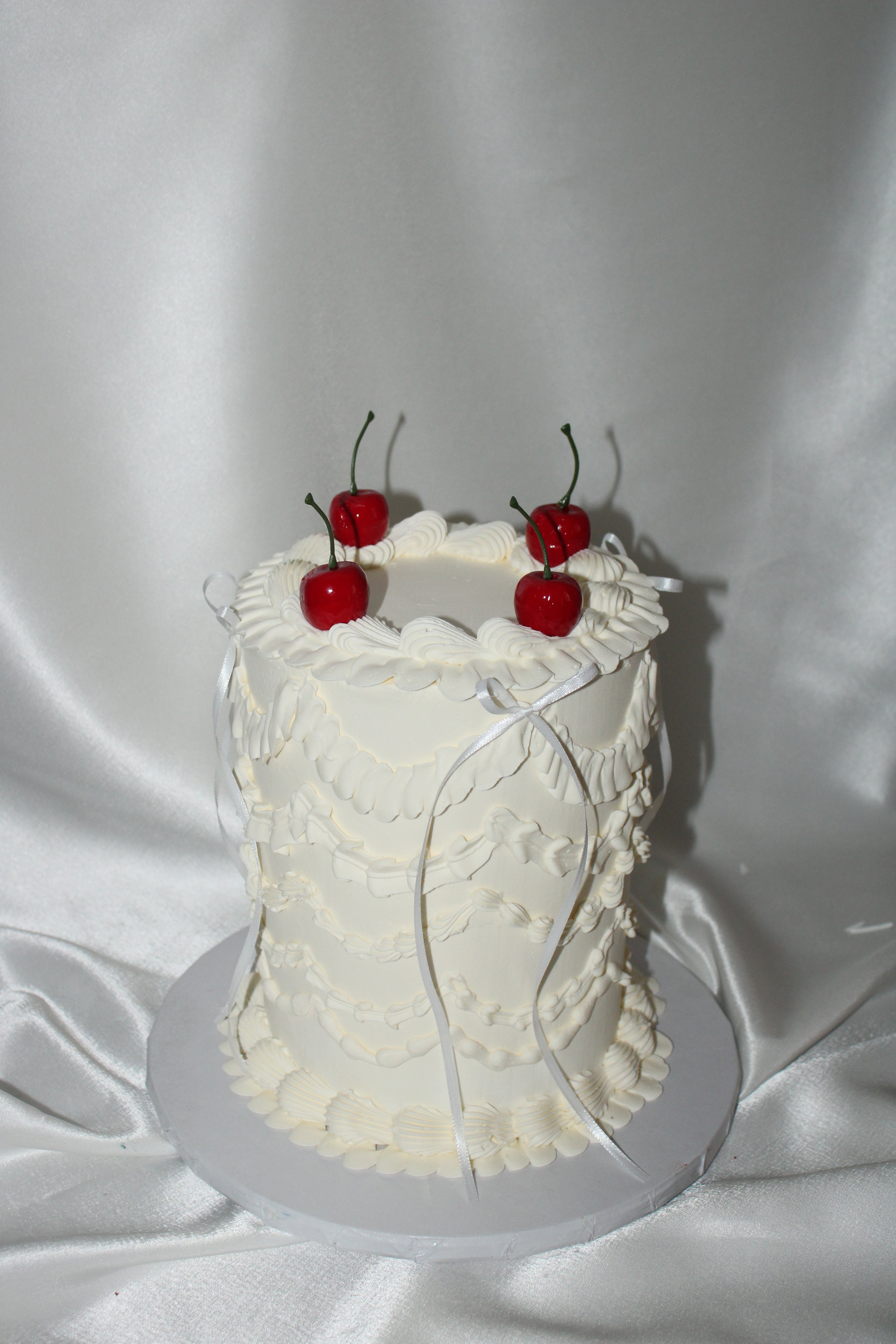 A round white frosted cake decorated with piped floral patterns and topped with four red cherries, presented on a white round board against a draped white satin fabric background.