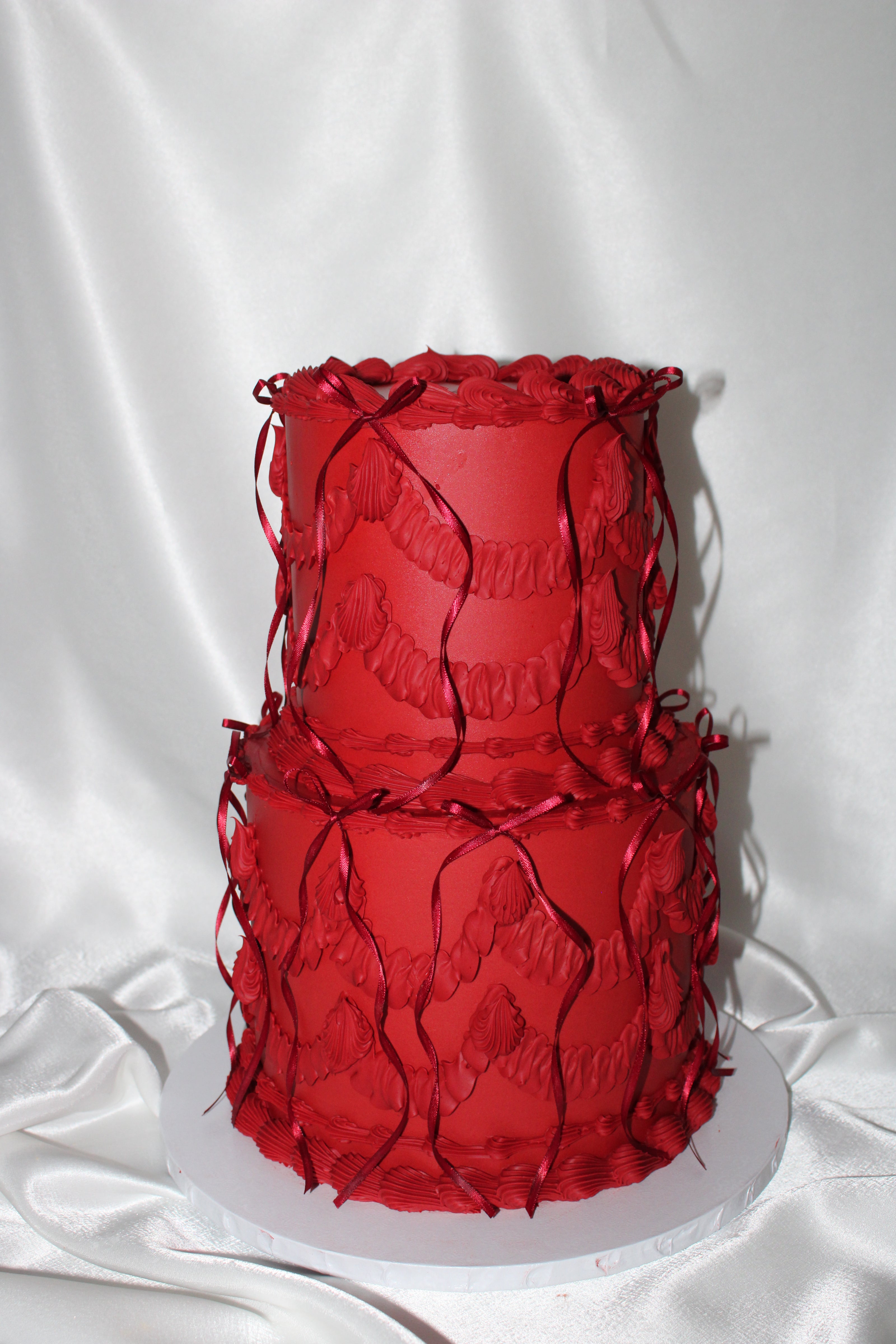A two-tier red cake decorated with textured ruffles and hanging red ribbons, displayed on a white round cake board against a white satin fabric backdrop.