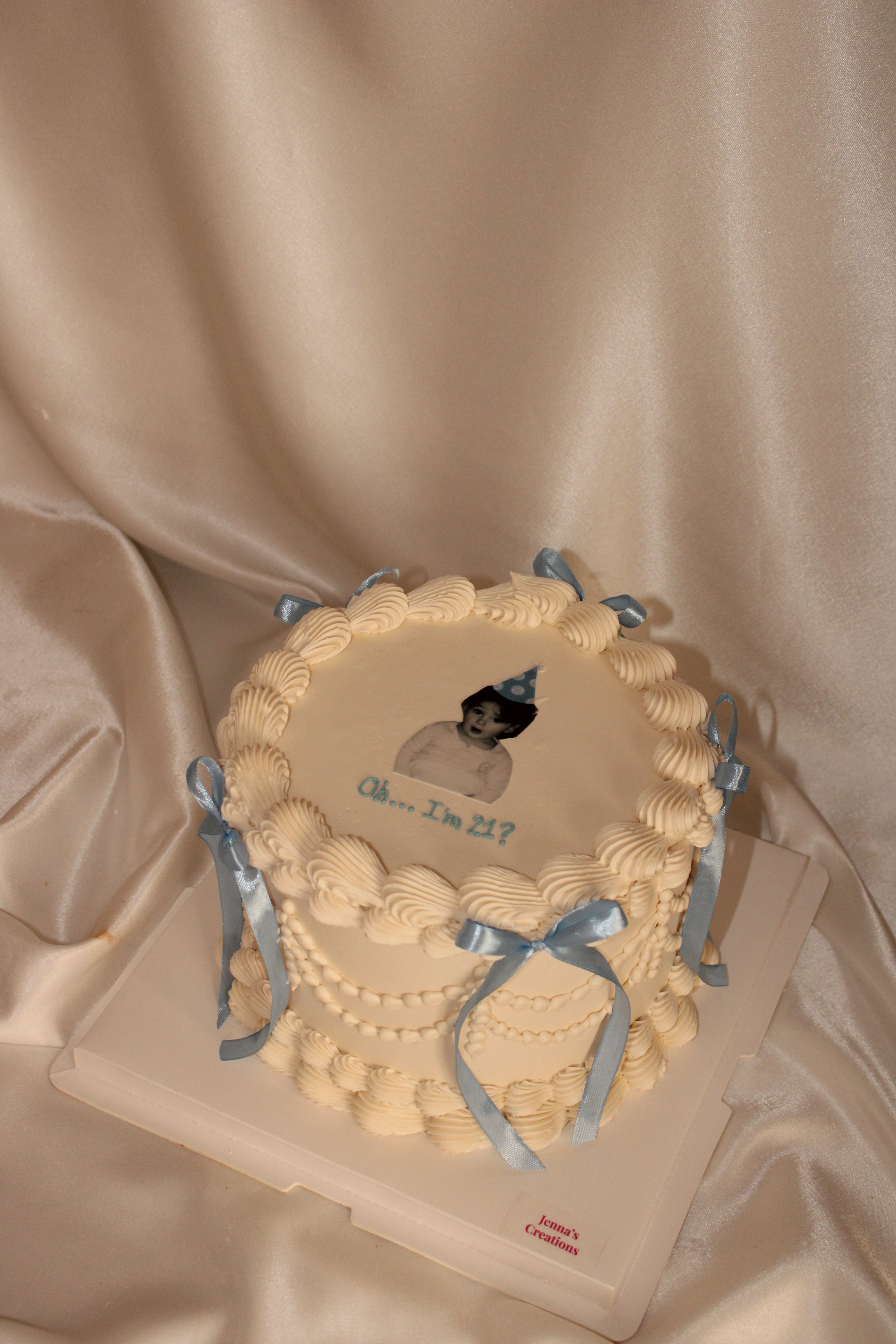 A round birthday cake with cream-colored frosting, piped borders, and light blue ribbon bows, decorated with a custom portrait and the text 'Oh my, I'm 21?' on top, set on a white cake board against a draped satin fabric background.