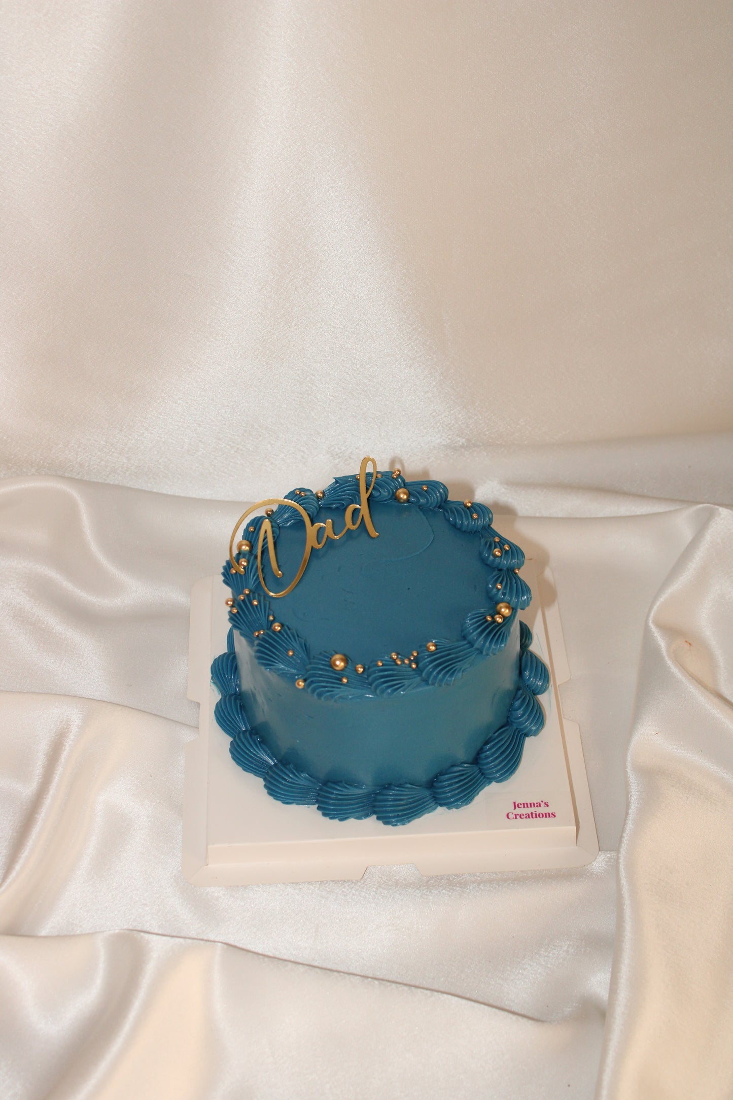 Royal blue buttercream cake with gold “Dad” topper and gold pearl decorations on a white cake board.
