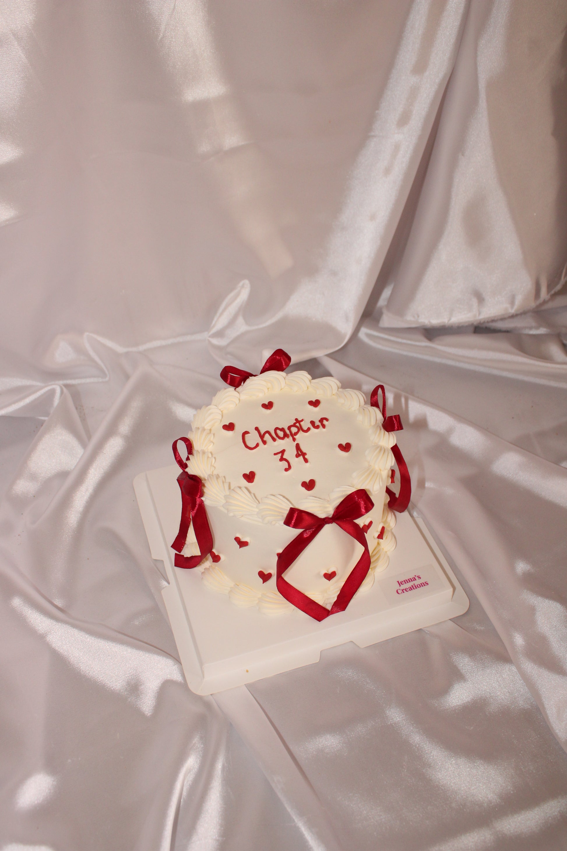 A small white frosted cake decorated with red ribbon bows and tiny red hearts, with “Chapter 34” written on top, displayed on a satin fabric background.