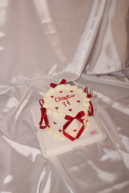A small white frosted cake decorated with red ribbon bows and tiny red hearts, with “Chapter 34” written on top, displayed on a satin fabric background.