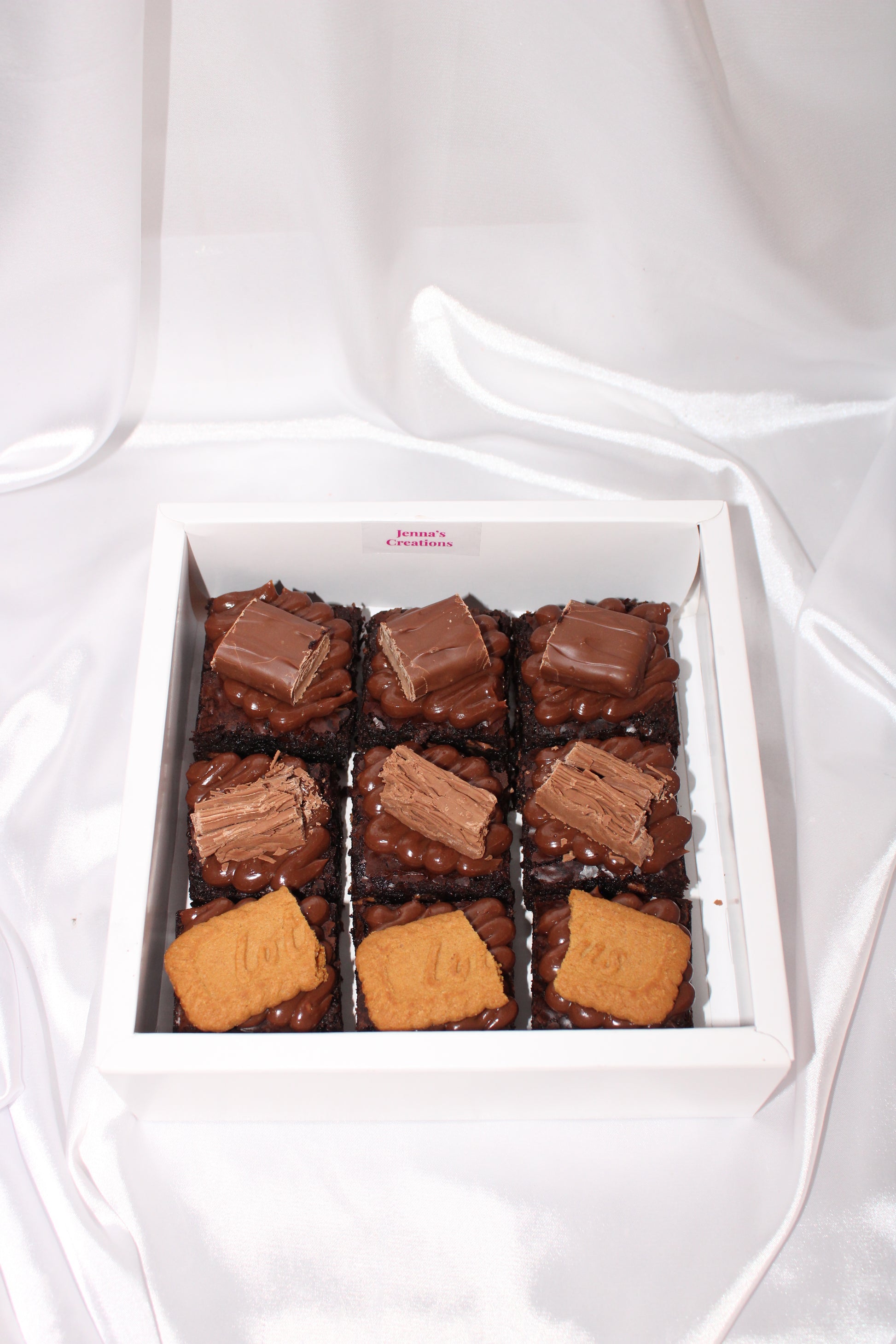 Assorted chocolate brownies topped with melted chocolate, biscuit pieces, and chocolate bars in a white box