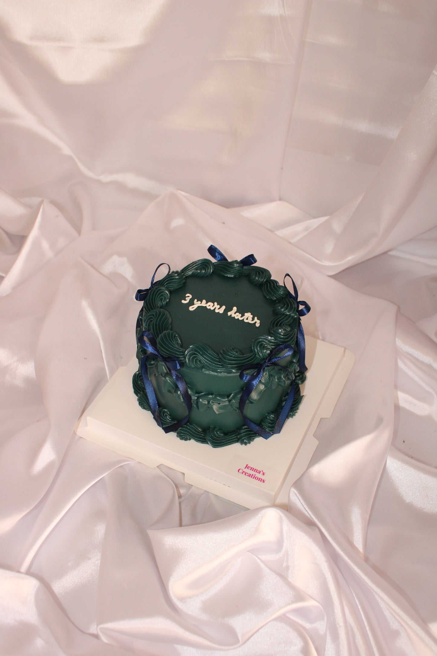 dark green vintage-style cake with blue ribbon bows and ‘3 years later’ written on top, displayed on a white satin backdrop