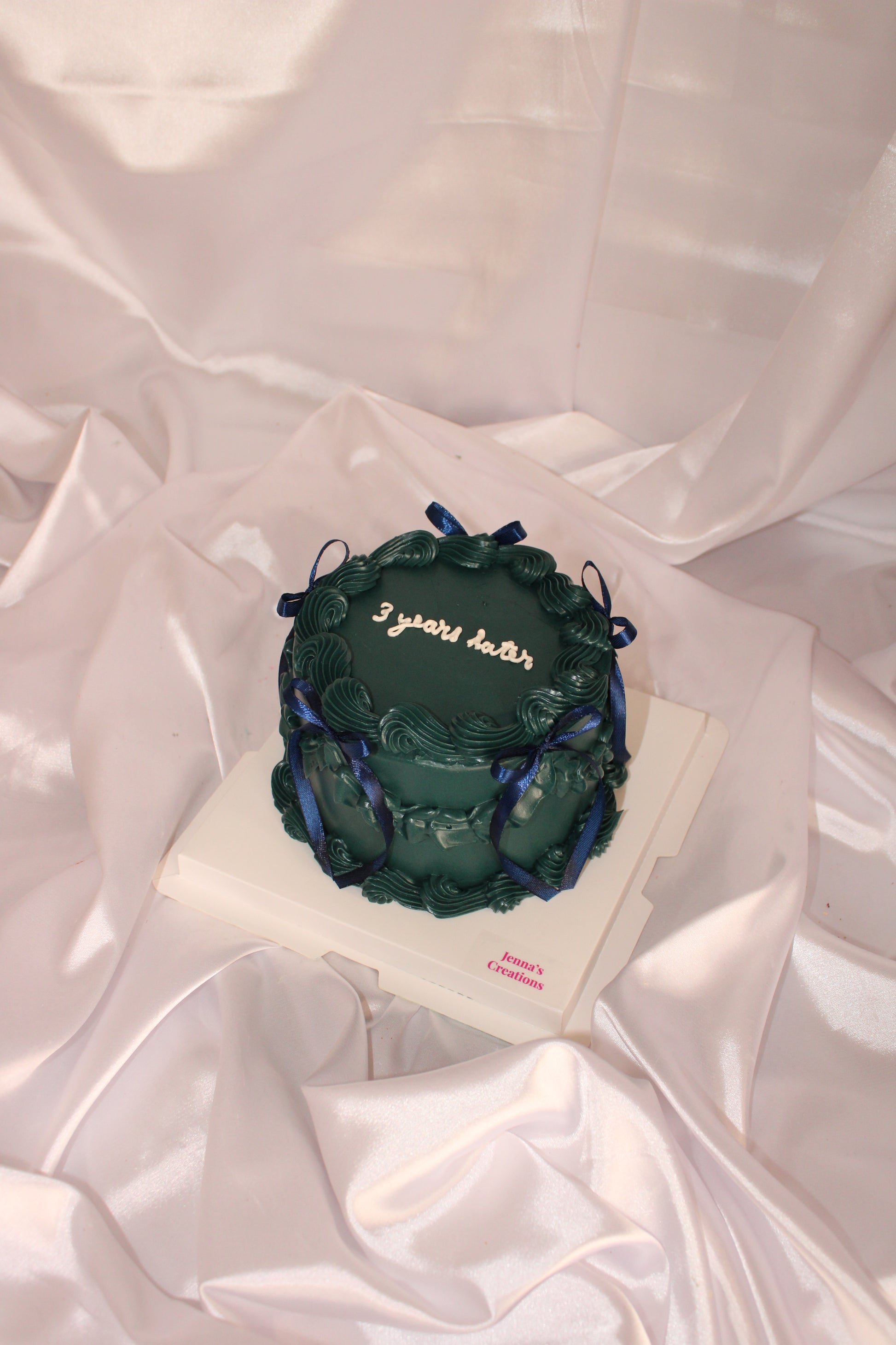 dark green vintage-style cake with blue ribbon bows and ‘3 years later’ written on top, displayed on a white satin backdrop