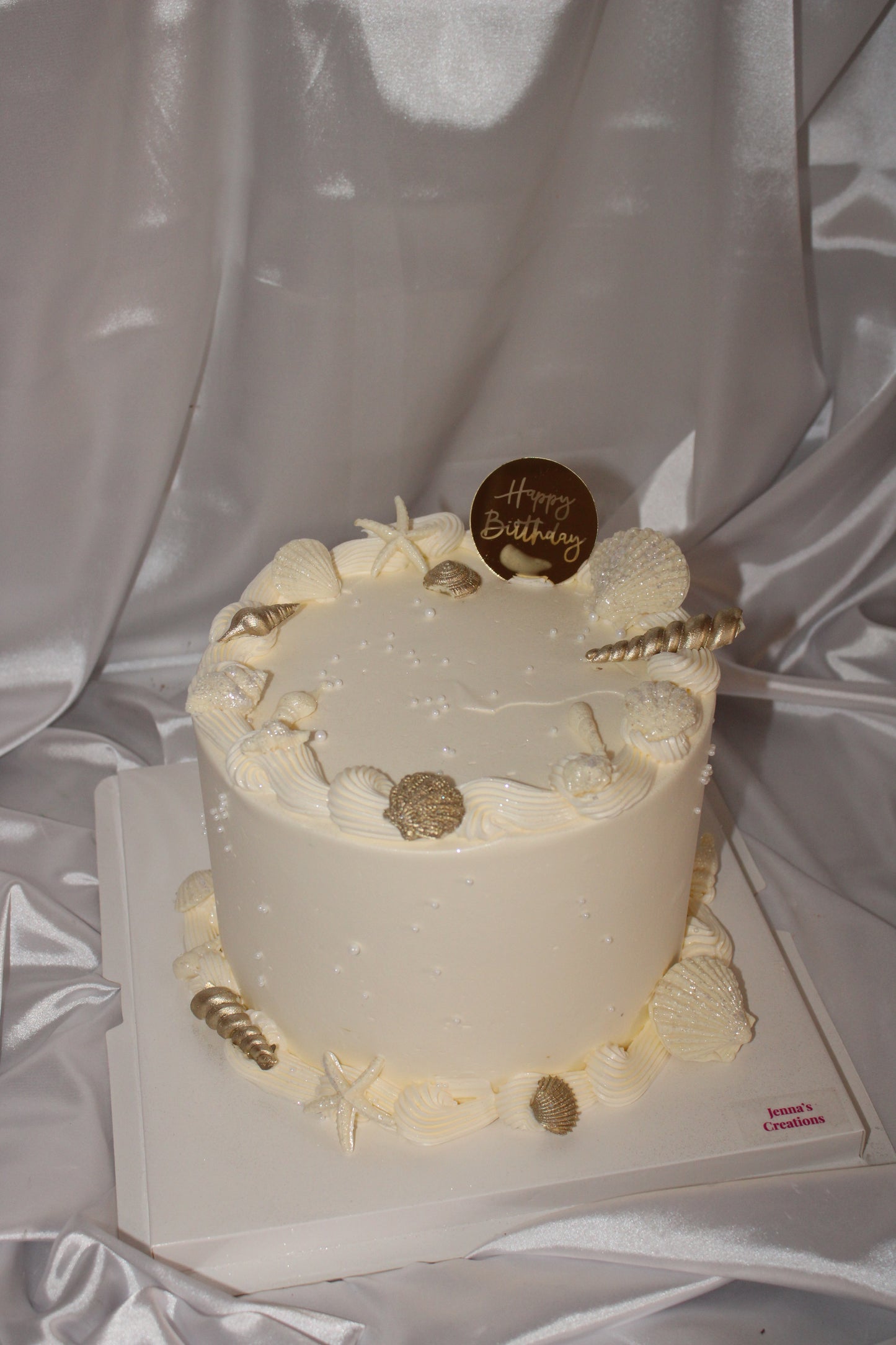 Ivory buttercream cake decorated with edible seashells, gold accents, and pearl details on a satin backdrop.