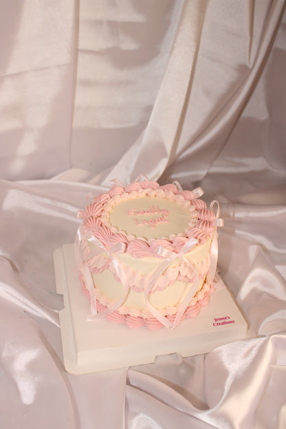 Vintage-style cream and pink cake with ribbon bows and a personalized name on top, displayed on a white satin backdrop.