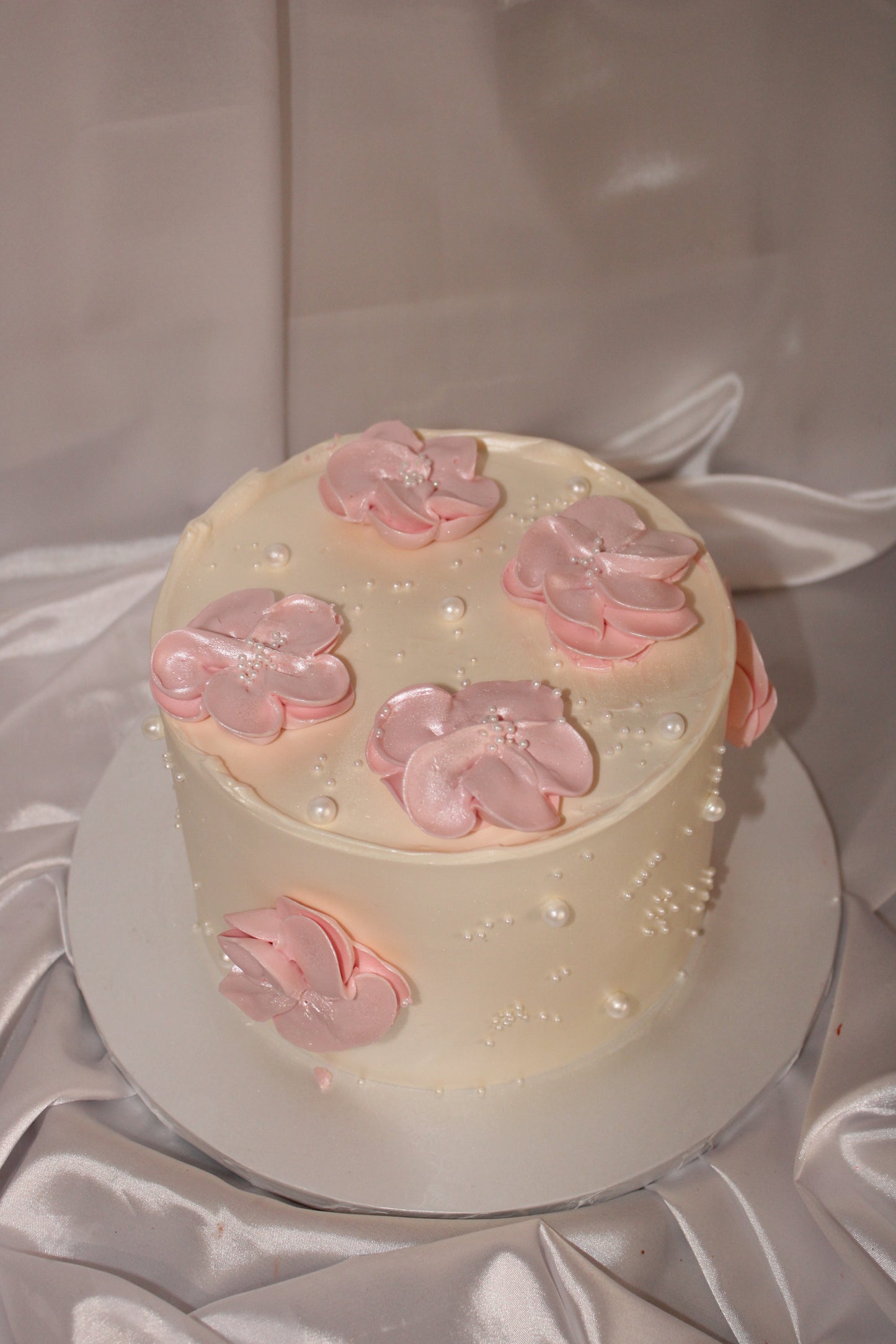 Elegant ivory buttercream cake decorated with soft pink floral accents and pearl details on a satin backdrop.